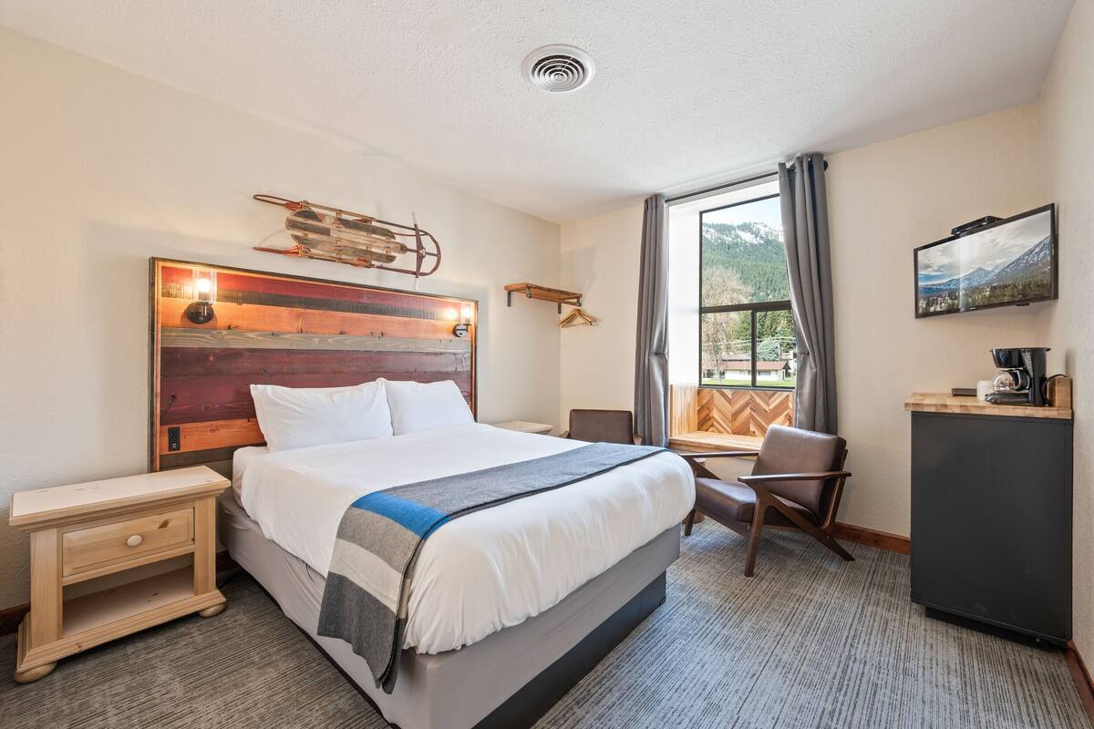 Quarto de hotel com uma cama grande, uma escrivaninha, uma cadeira, um armário pequeno e uma janela com vista para montanhas.