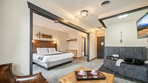 A hotel room with a bed, a wooden coffee table with breakfast items, a seating area with a leather chair, a kitchen area with a black bathtub, and a mounted TV on the wall. - Hotel Leavenworth (Leavenworth)