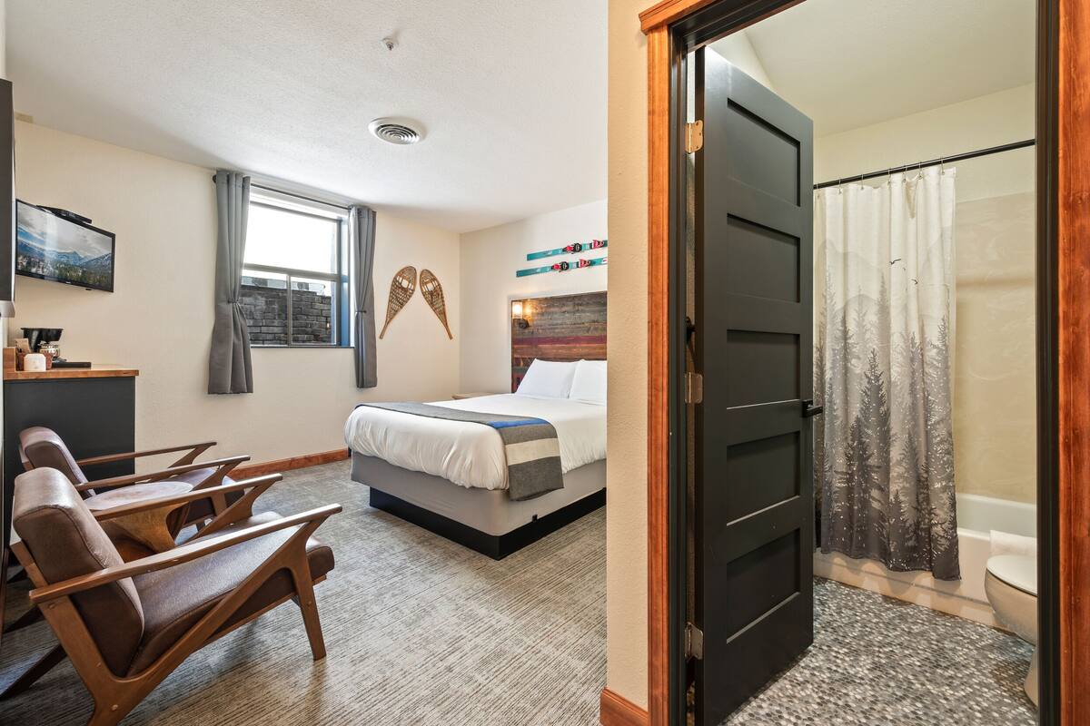 Quarto de hotel com cama, escrivaninha, cadeira, janela com cortinas, cortina do chuveiro e porta.