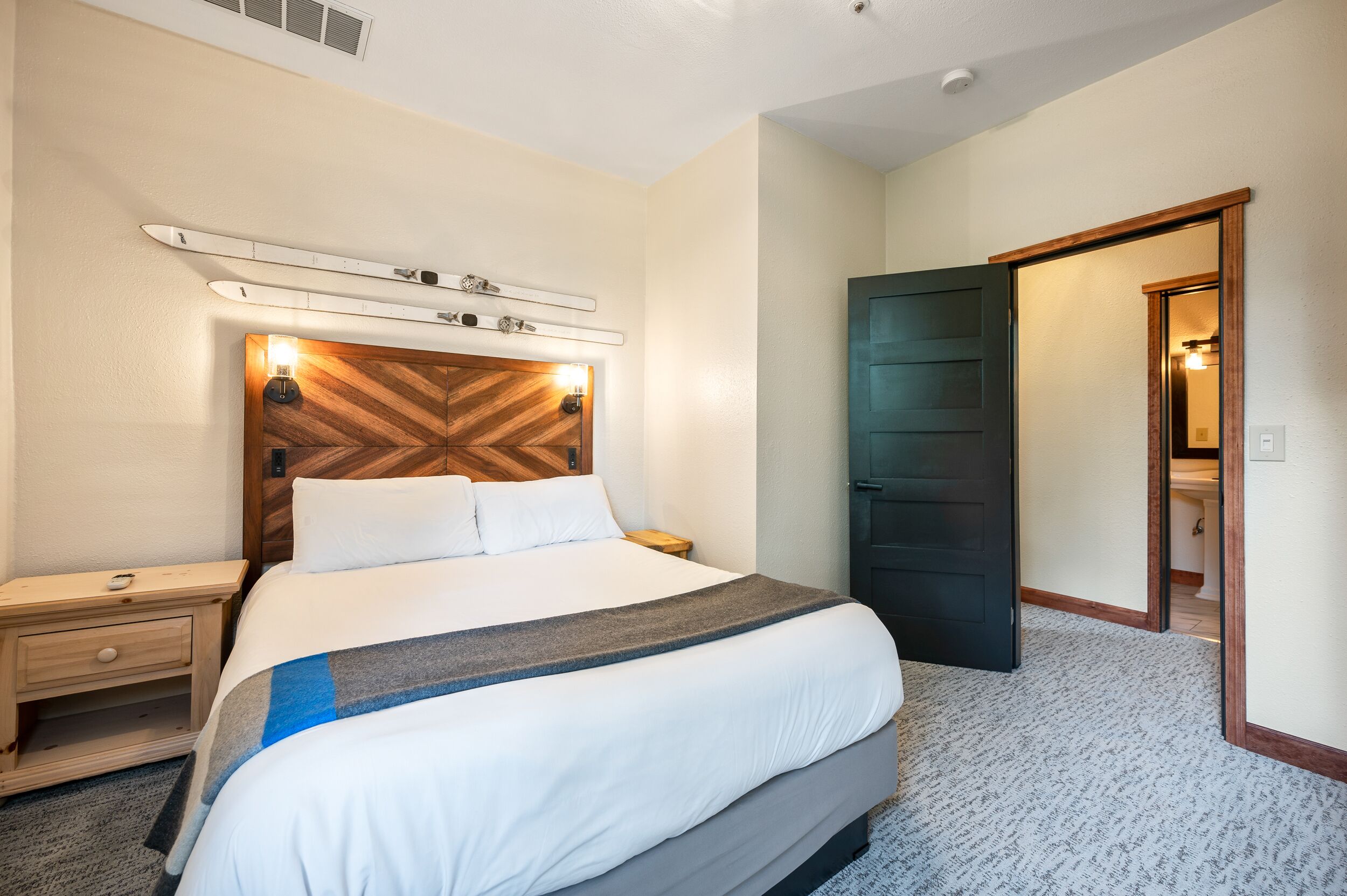 A hotel room with a large bed, a wooden headboard, a nightstand, a desk, and a door leading to a bathroom.