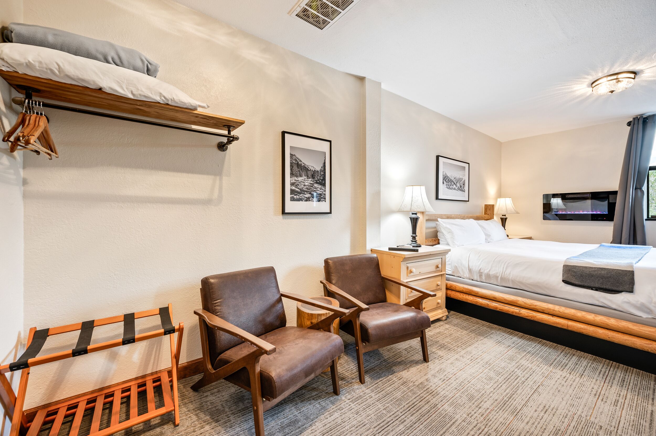 A hotel room with a bed, two chairs, a wooden shelf with pillows, a nightstand with a lamp, and a TV.