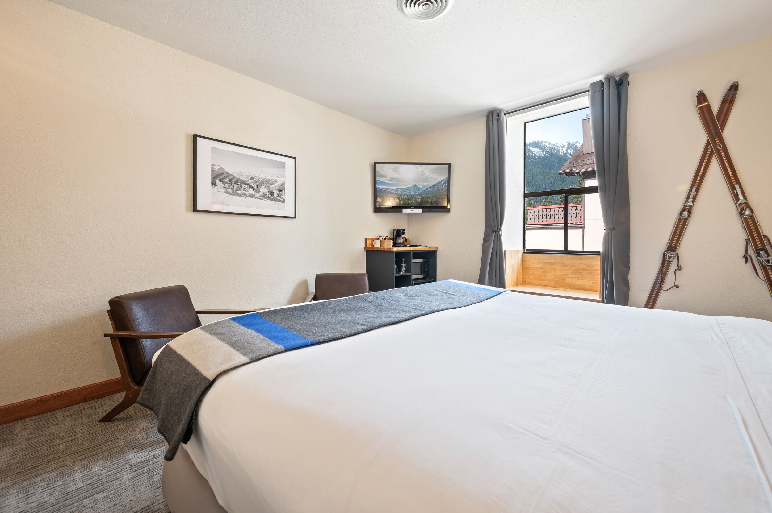 Una habitación de hotel con una cama grande, un escritorio, una silla, una mesita de noche con una lámpara, y una ventana con vistas a montañas.