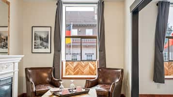 Habitación de hotel con chimenea, dos sillones de cuero, una mesa de centro de madera y un ventanal con cortinas.