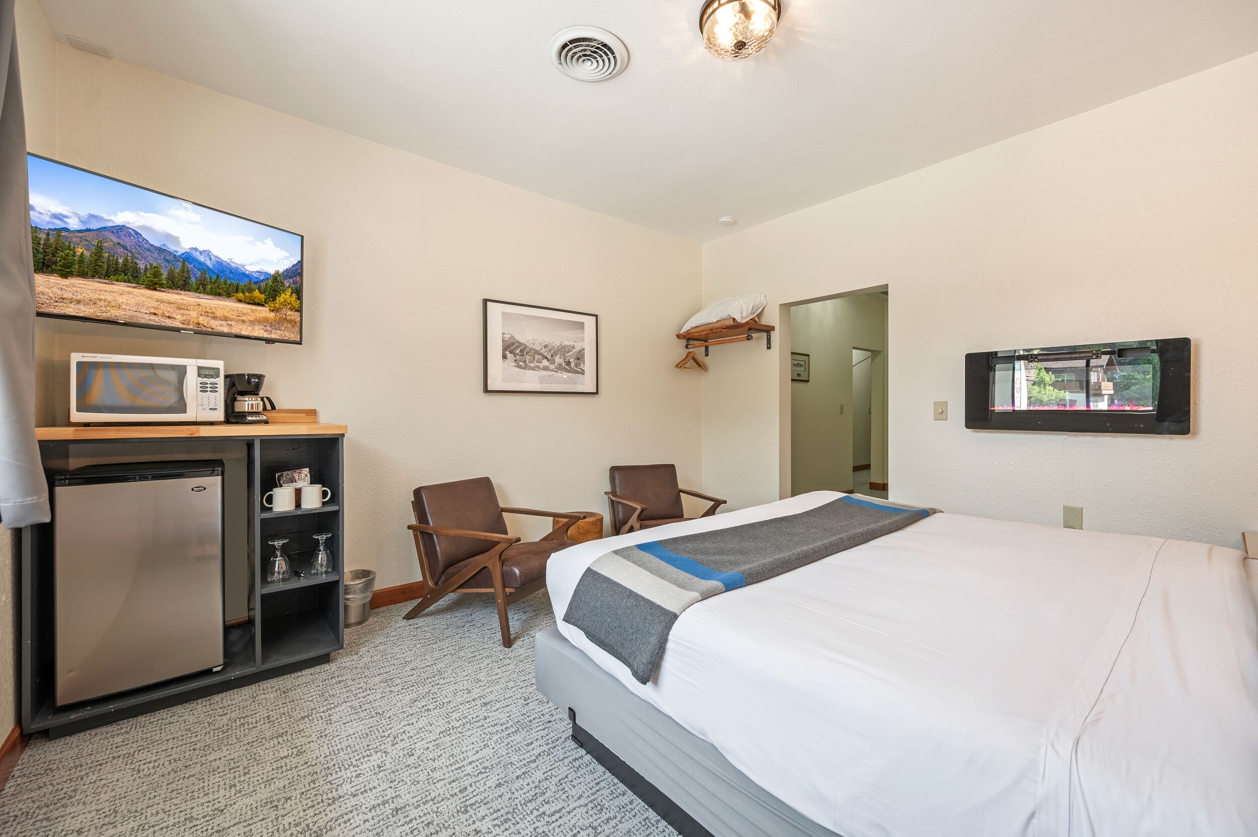 A hotel room with a bed, a desk with a microwave, a refrigerator, two chairs, a TV, and a framed picture on the wall.