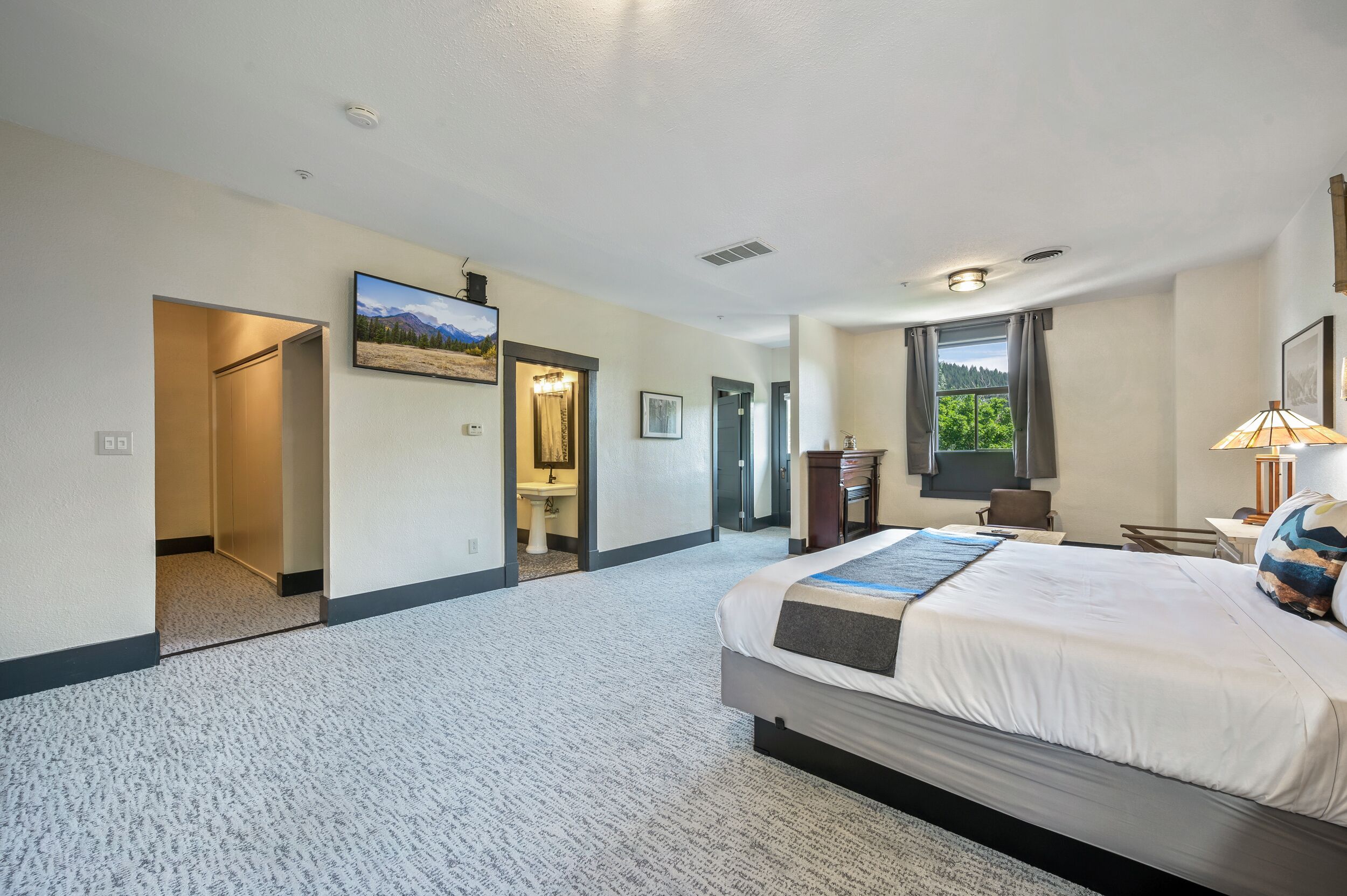 Ein Hotelzimmer mit einem großen Bett, einem Schreibtisch, einem Stuhl, einem Fernseher und einem Fenster mit Blick auf Bäume.