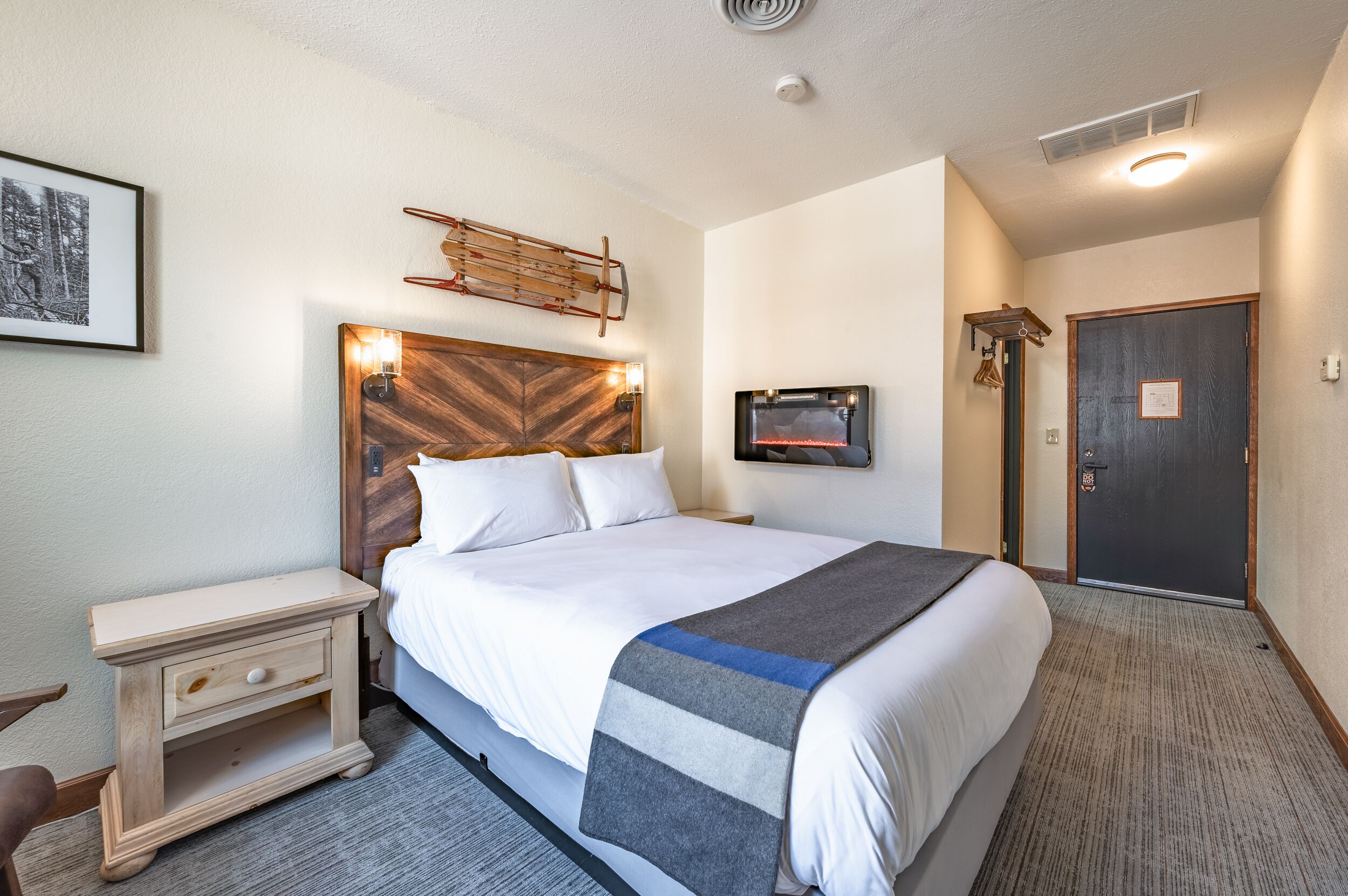 Quarto de hotel com uma cama grande, uma mesinha de cabeceira, televisão na parede e uma porta.