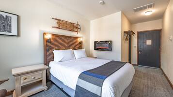 Quarto de hotel com uma cama grande, uma mesinha de cabeceira, televisão na parede e uma porta.