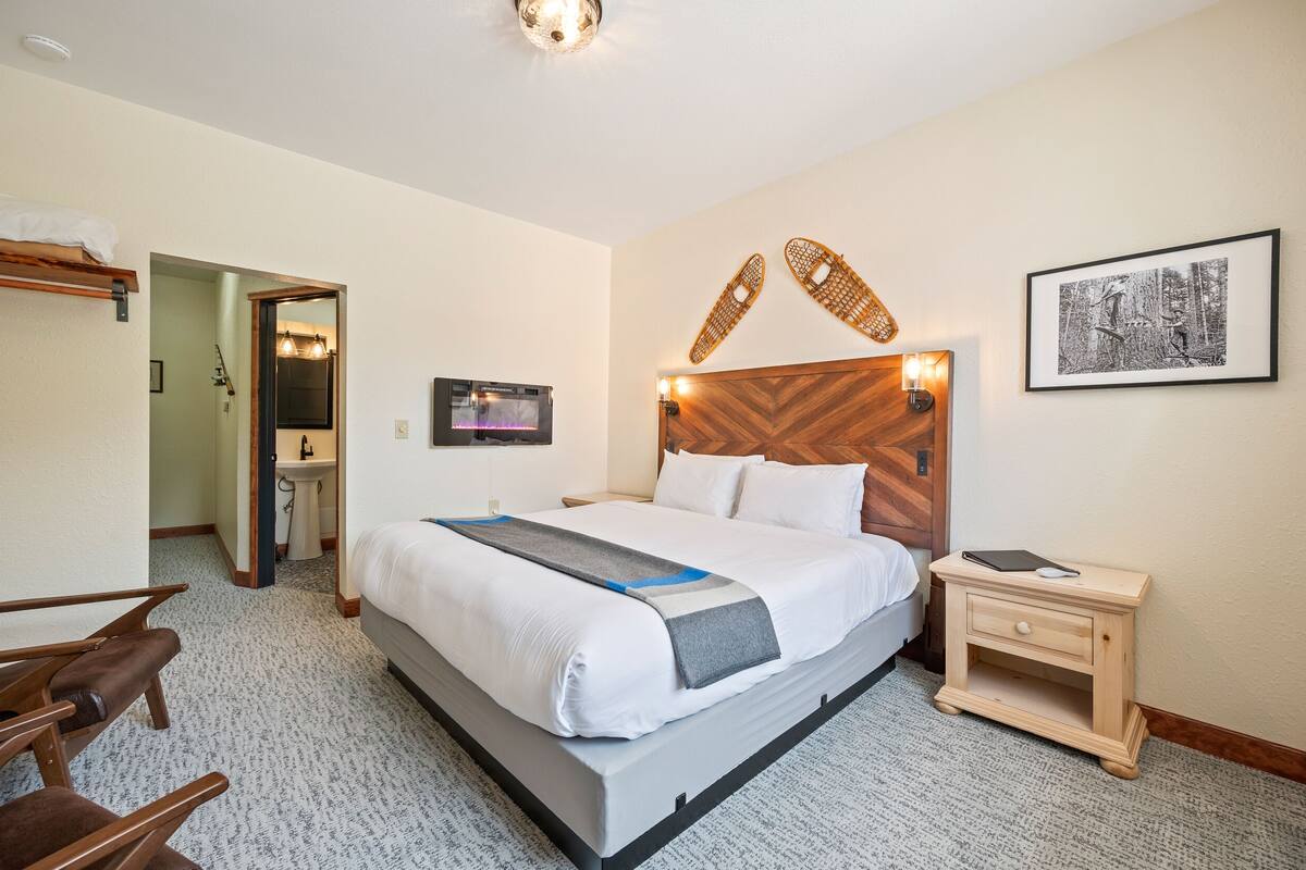 Quarto de hotel com cama, mesa de cabeceira, cadeira e banheiro visível por uma porta aberta.