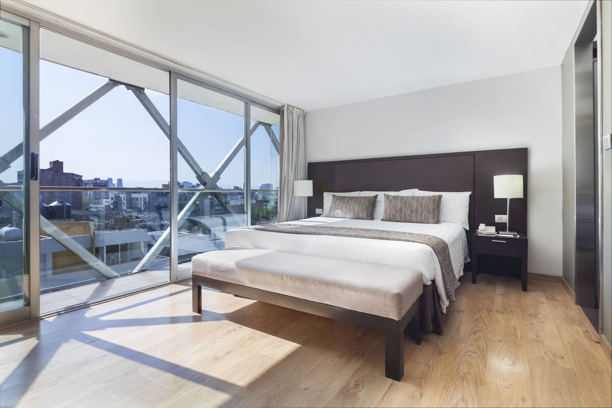 A modern hotel room with a large bed, bedside tables, and a view of the city through floor-to-ceiling windows.