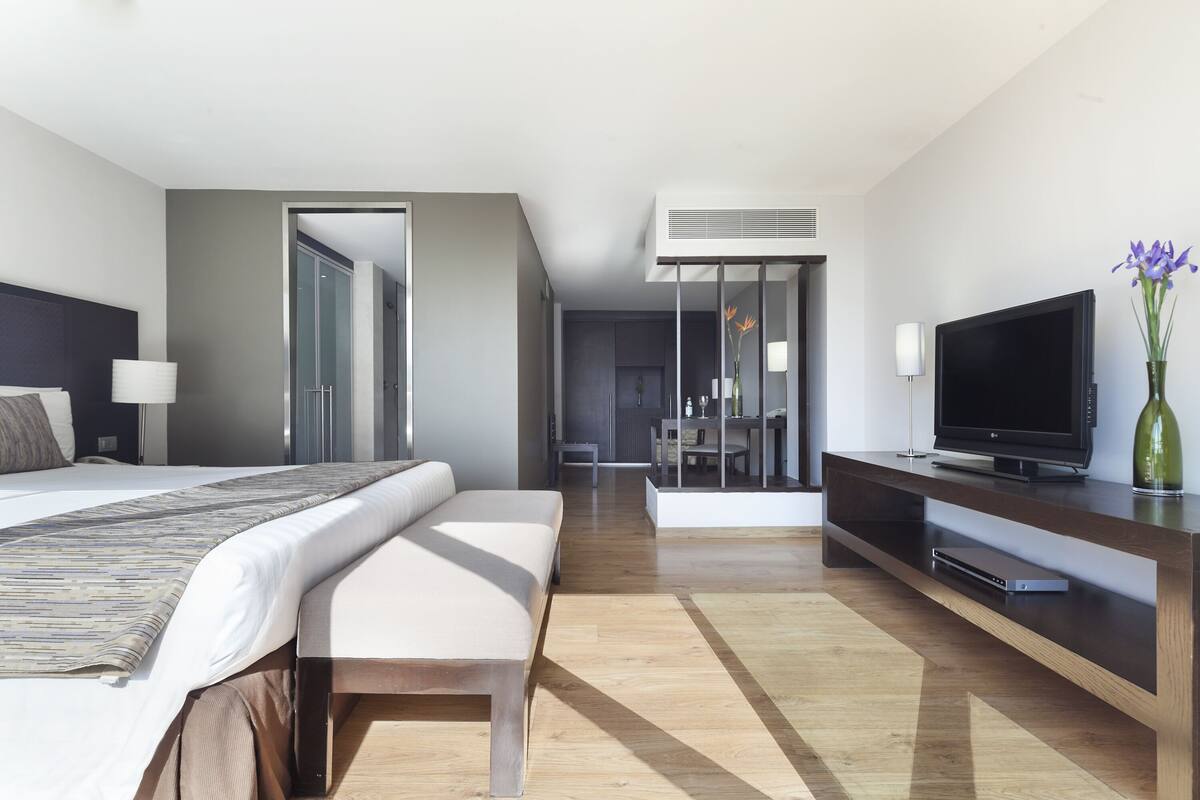 A modern hotel room with a large bed, a TV, a desk, and a dining area.