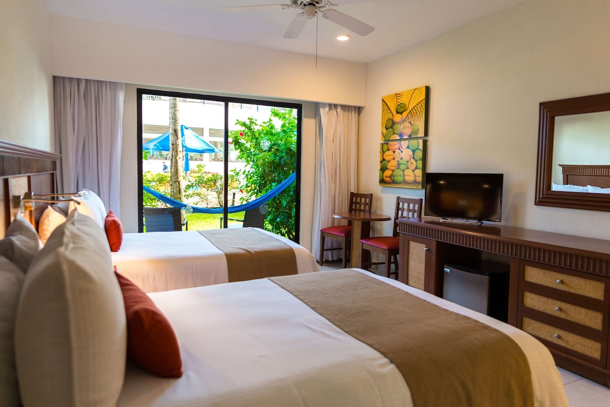 Habitación de hotel con dos camas, televisión, escritorio y vista a un jardín con una hamaca.