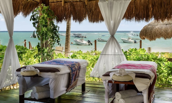 A beachside room with two beds, a thatched roof, and a view of boats on the water.