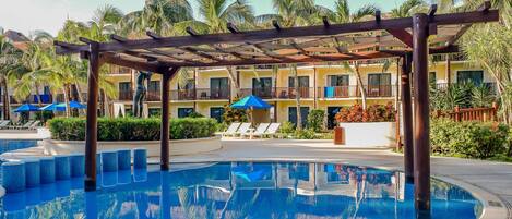 A pool area with lounge chairs and umbrellas, surrounded by palm trees and a multi-story building.
