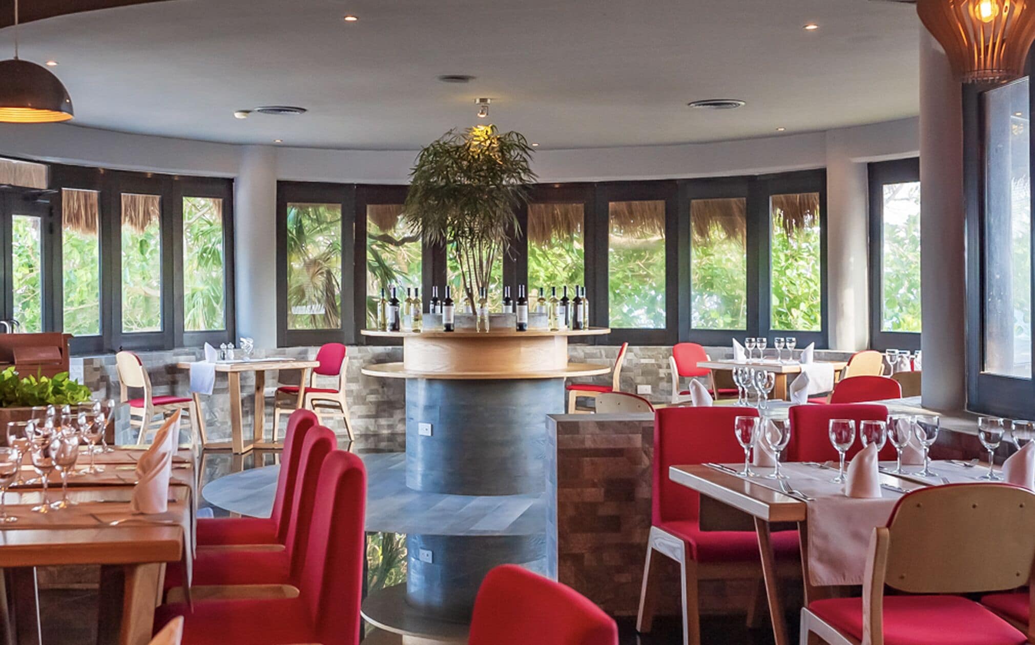 A restaurant with round tables, red chairs, a central bar, and large windows with greenery outside.