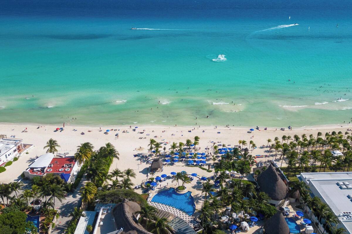 Vue aérienne d’un complexe hôtelier en bord de mer, avec une eau turquoise cristalline, des palmiers et un espace piscine.