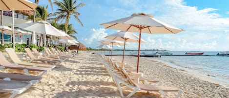 A beach with rows of sun loungers and umbrellas, palm trees, and boats in the background.