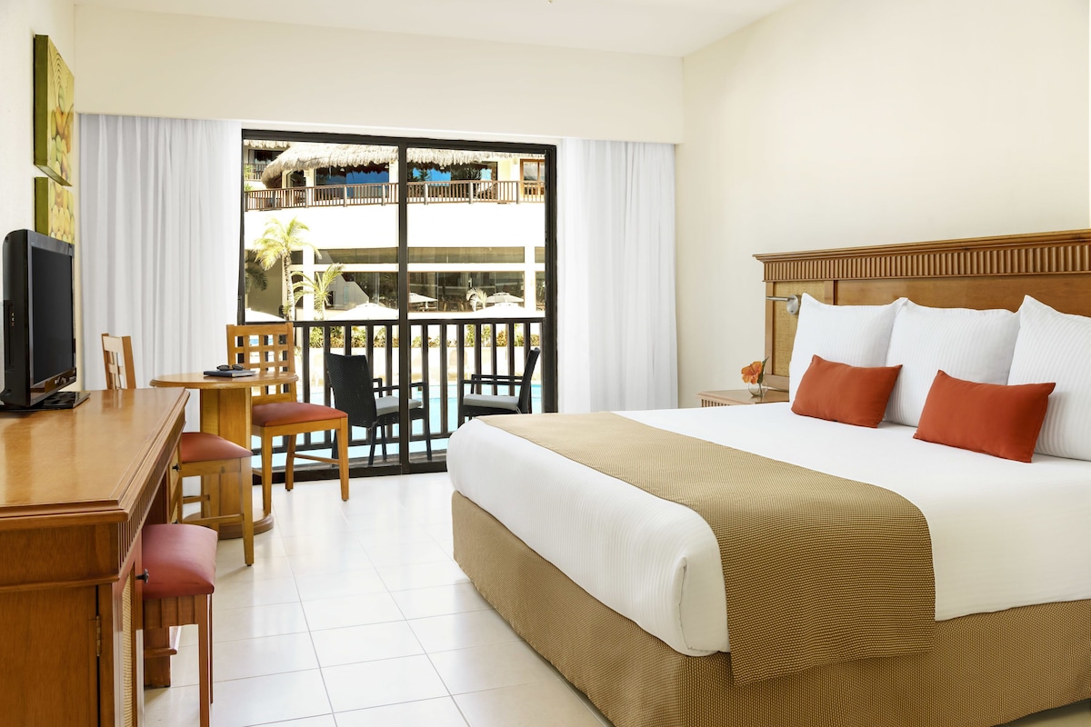 Habitación de hotel con una cama grande, un escritorio con silla, televisor y balcón con vista a una piscina y edificios.