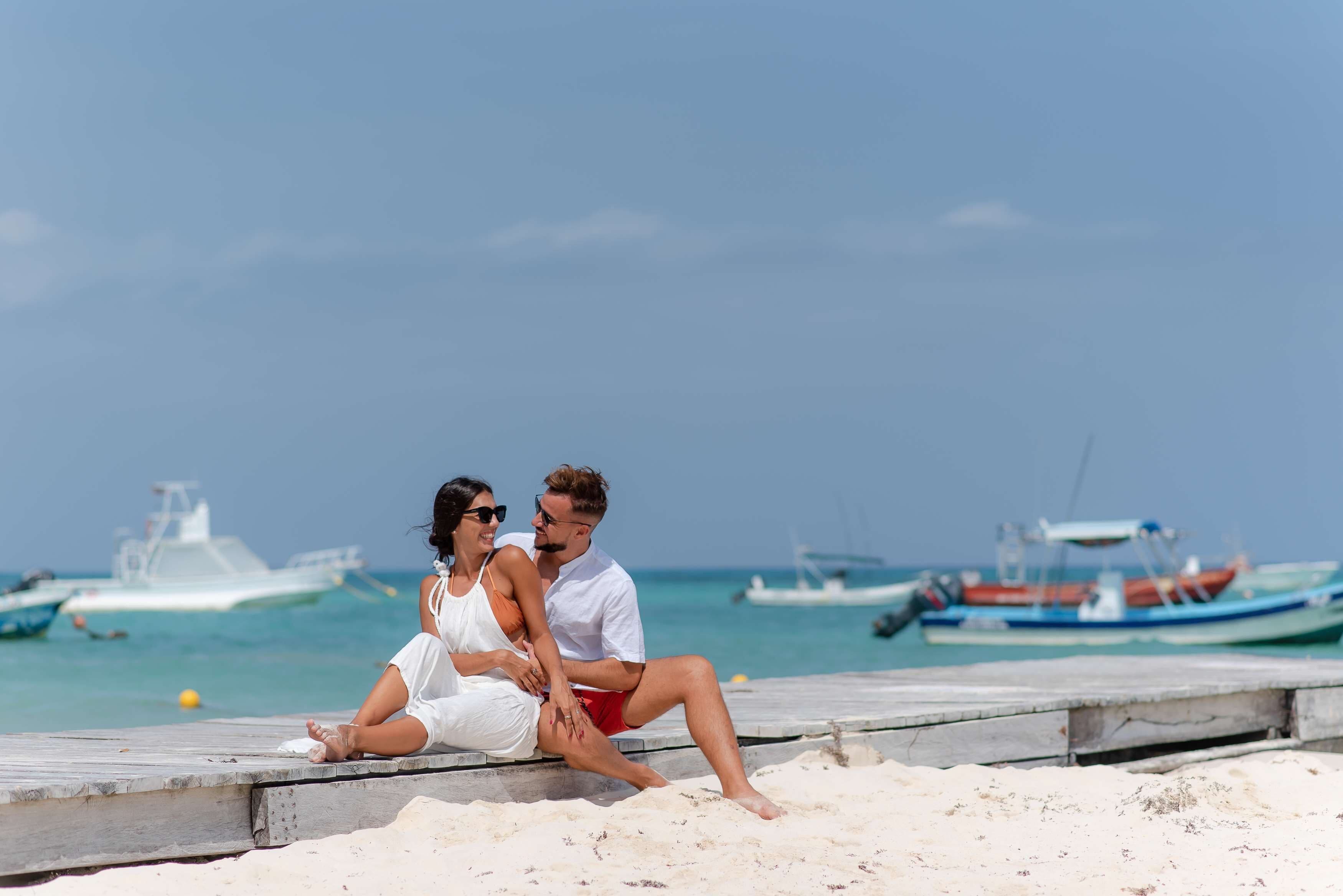 A couple sitting on a wooden pier by the beach with boats in the background.