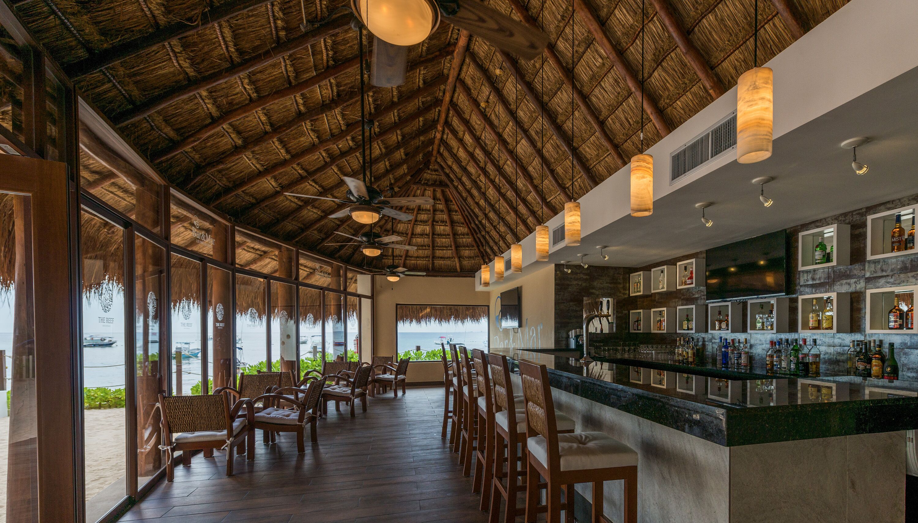 A bar area with a view of the sea, wooden chairs, and a thatched roof ceiling.