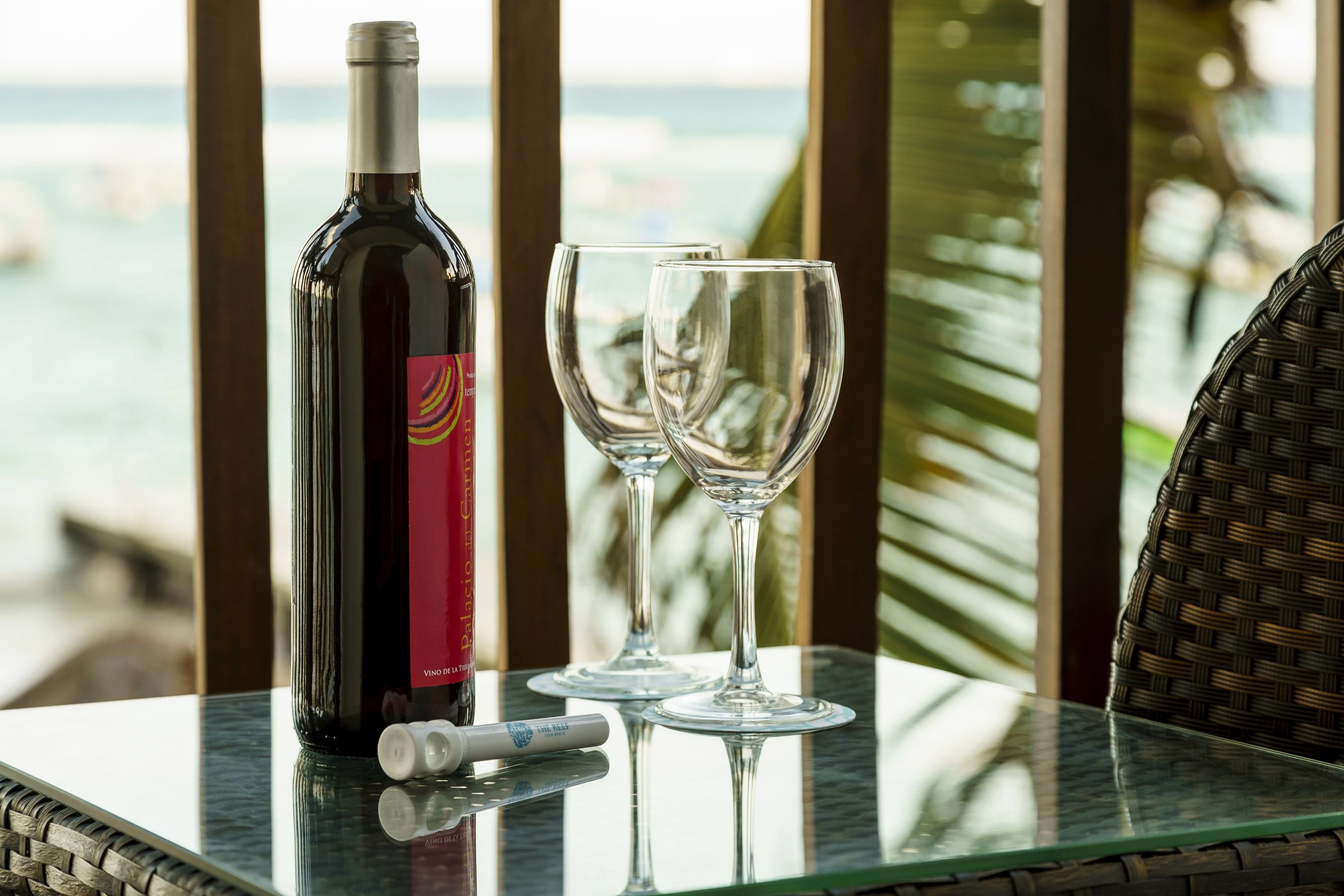 A table with a bottle of wine and two glasses on a glass table near a window overlooking the ocean.