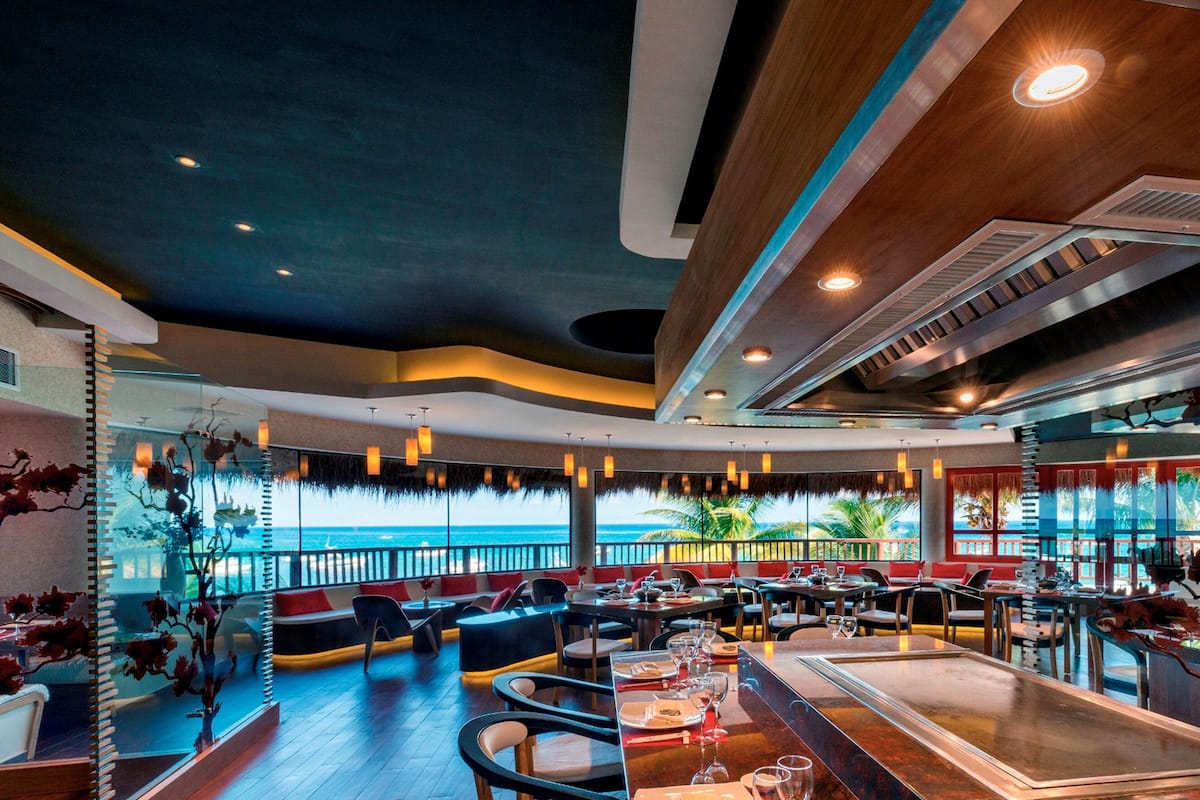 Un restaurant moderne offrant une vue sur l’océan, avec des tables en bois, des sièges rembourrés et un éclairage suspendu.