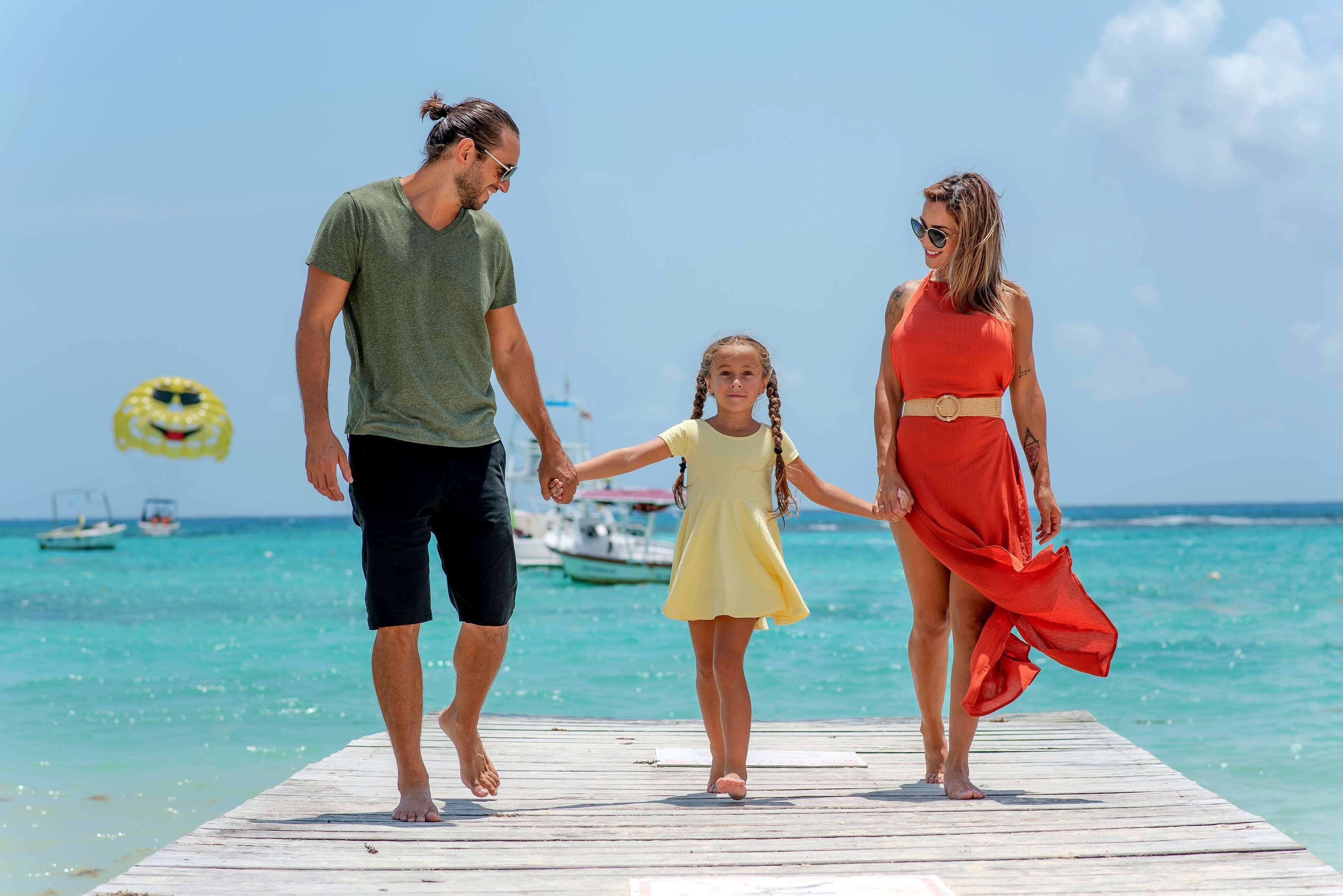 Una familia caminando sobre un muelle de madera, con un cielo azul despejado y un mar tranquilo al fondo.