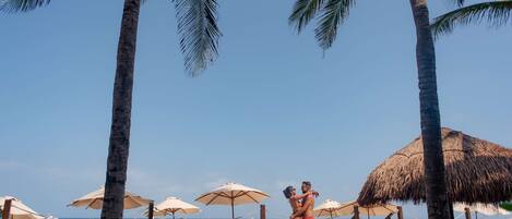 A beach with palm trees, umbrellas, and a couple embracing.
