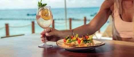 A dining table with a plate of food and a glass of drink, with a view of the ocean in the background.