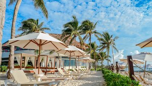 Ein Hotel am Strand mit Reihen von Liegestühlen und Sonnenschirmen, Palmen und blauem Himmel.