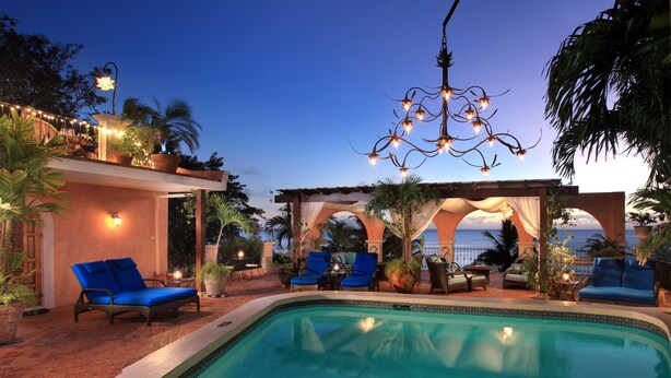 Una zona junto a la piscina con sillones reclinables azules, una pérgola con telas colgantes y un aplique.