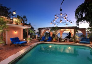 Ein Poolbereich mit blauen Liegestühlen, einer Pergola mit Vorhängen und einer hängenden Leuchte.