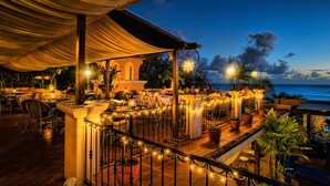 Un área de comedor al aire libre con mesas y sillas, luces colgantes y vista al océano al anochecer.
