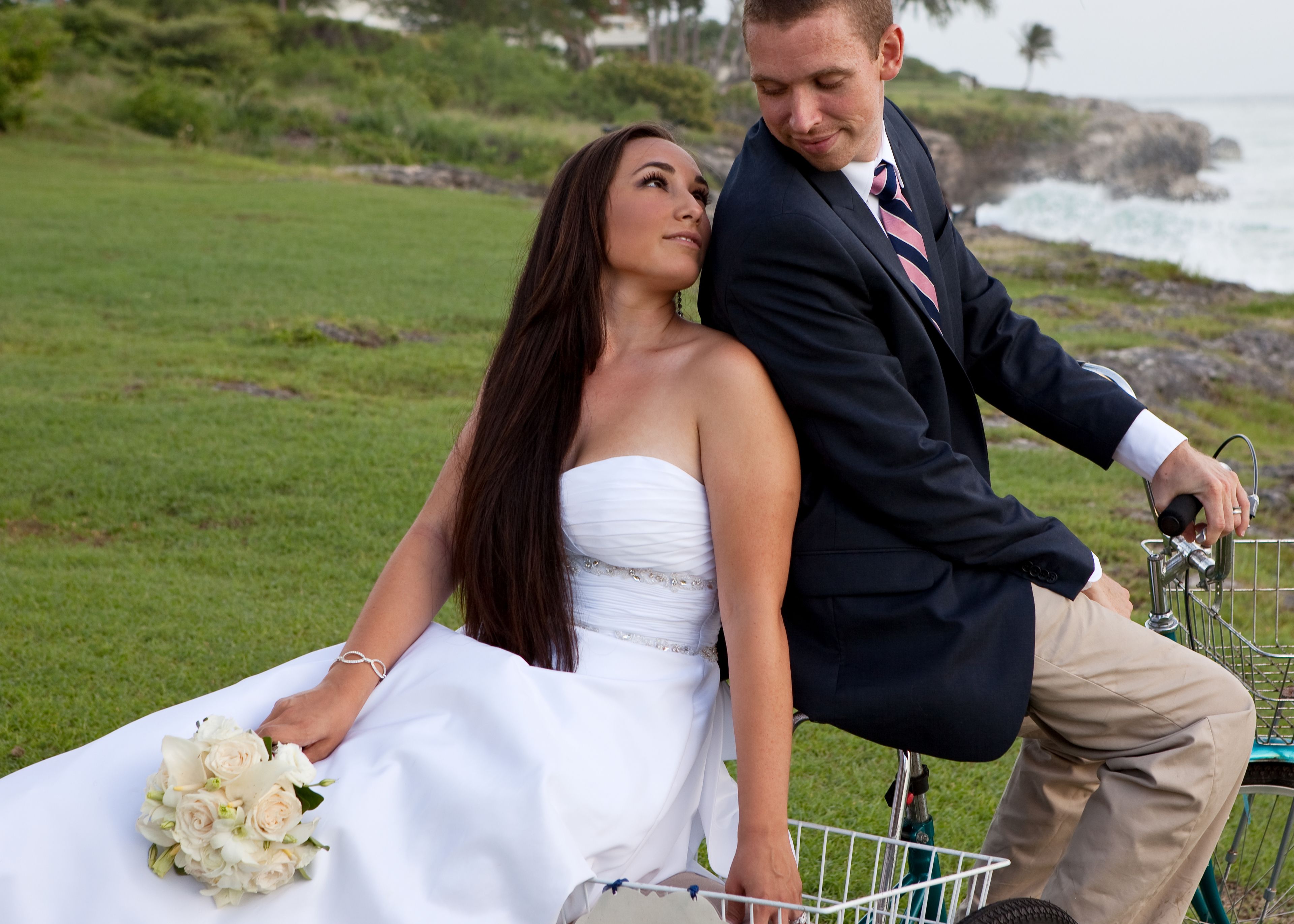 A couple on a bicycle in a grassy area with a view of the ocean.