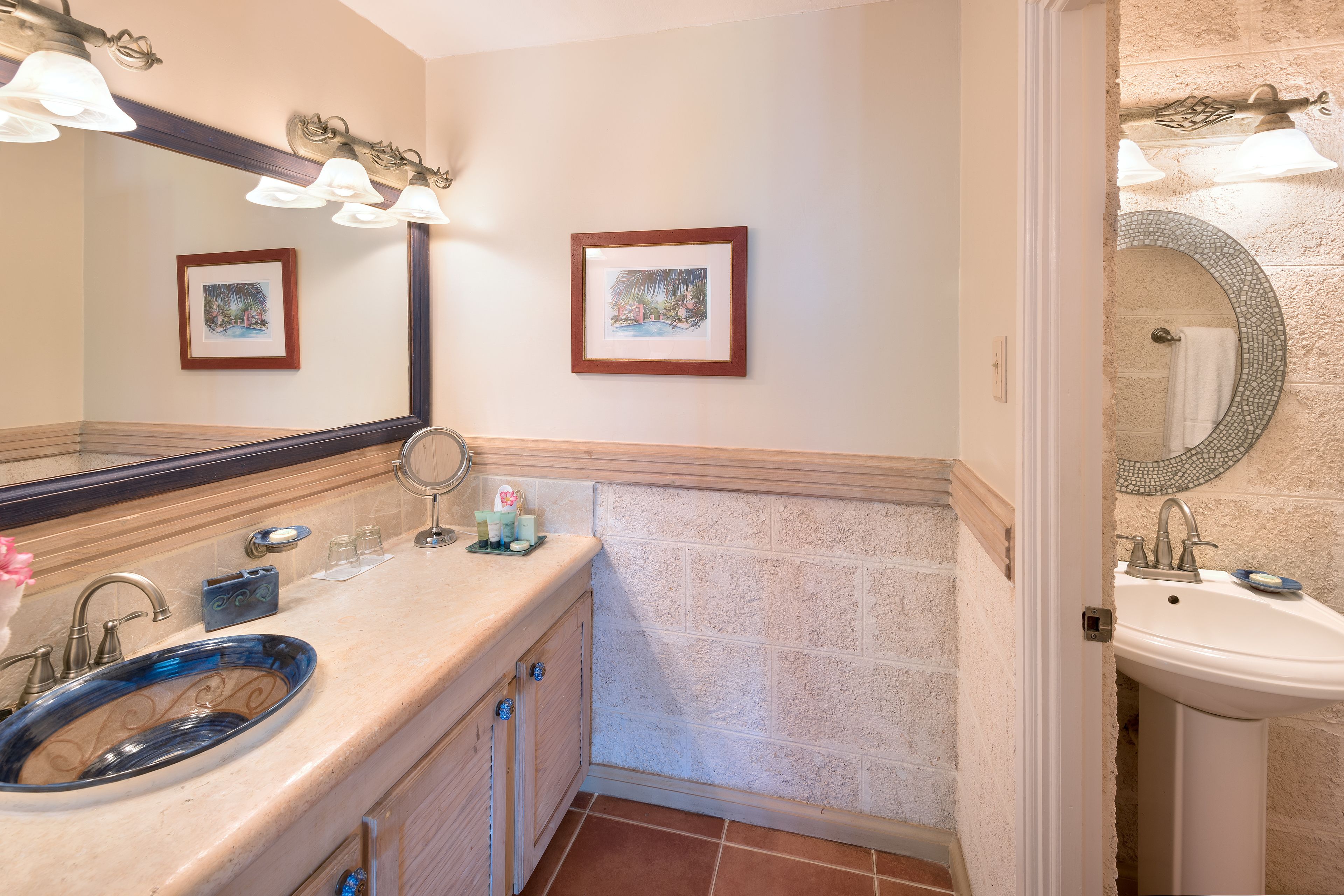 Un baño con un lavabo de vidrio azul, dos cuadros enmarcados en la pared y un lavabo blanco sobre pedestal.