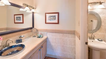 Un baño con un lavabo de vidrio azul, dos cuadros enmarcados en la pared y un lavabo blanco sobre pedestal.