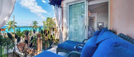 A balcony with blue cushioned seating, a glass door leading to an interior space, and a view of a pool and palm trees.