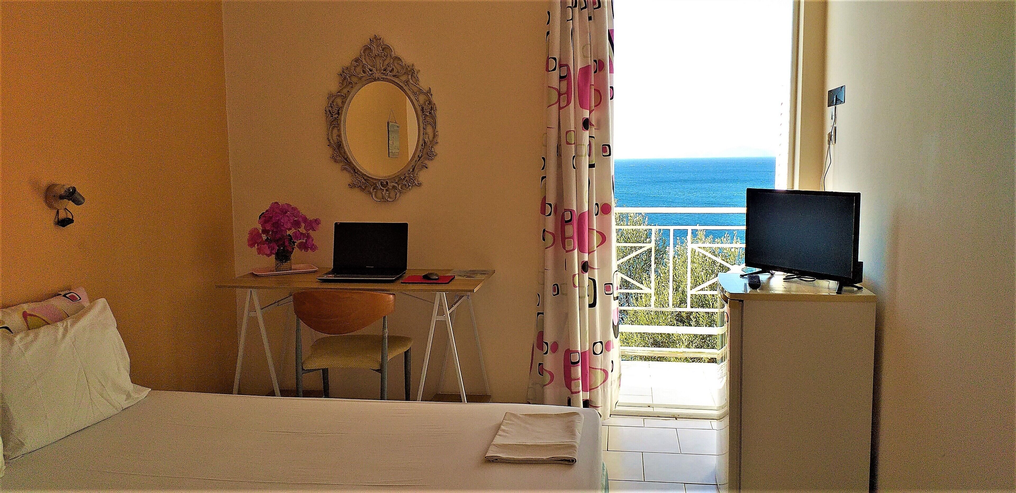 A hotel room with a bed, a desk with a laptop, a chair, a TV, and a view of the sea.
