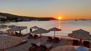 Ein Strand mit Liegestühlen und Strohdächern, ein Sonnenuntergang über dem Wasser und Boote in der Ferne.