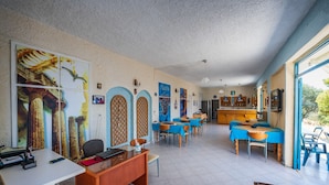 Ein geräumiger Essbereich mit blauen Tischen und Stühlen, ein Küchenbereich mit Holzschränken und ein großes Fenster, das für natürliches Licht sorgt.