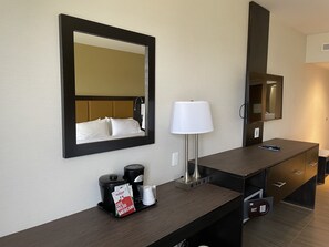 Quarto de hotel com um espelho grande, luminária de cabeceira, cafeteira e minibar.