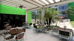 An outdoor patio area with tables and chairs, a ceiling fan, and a view of a building with palm trees.