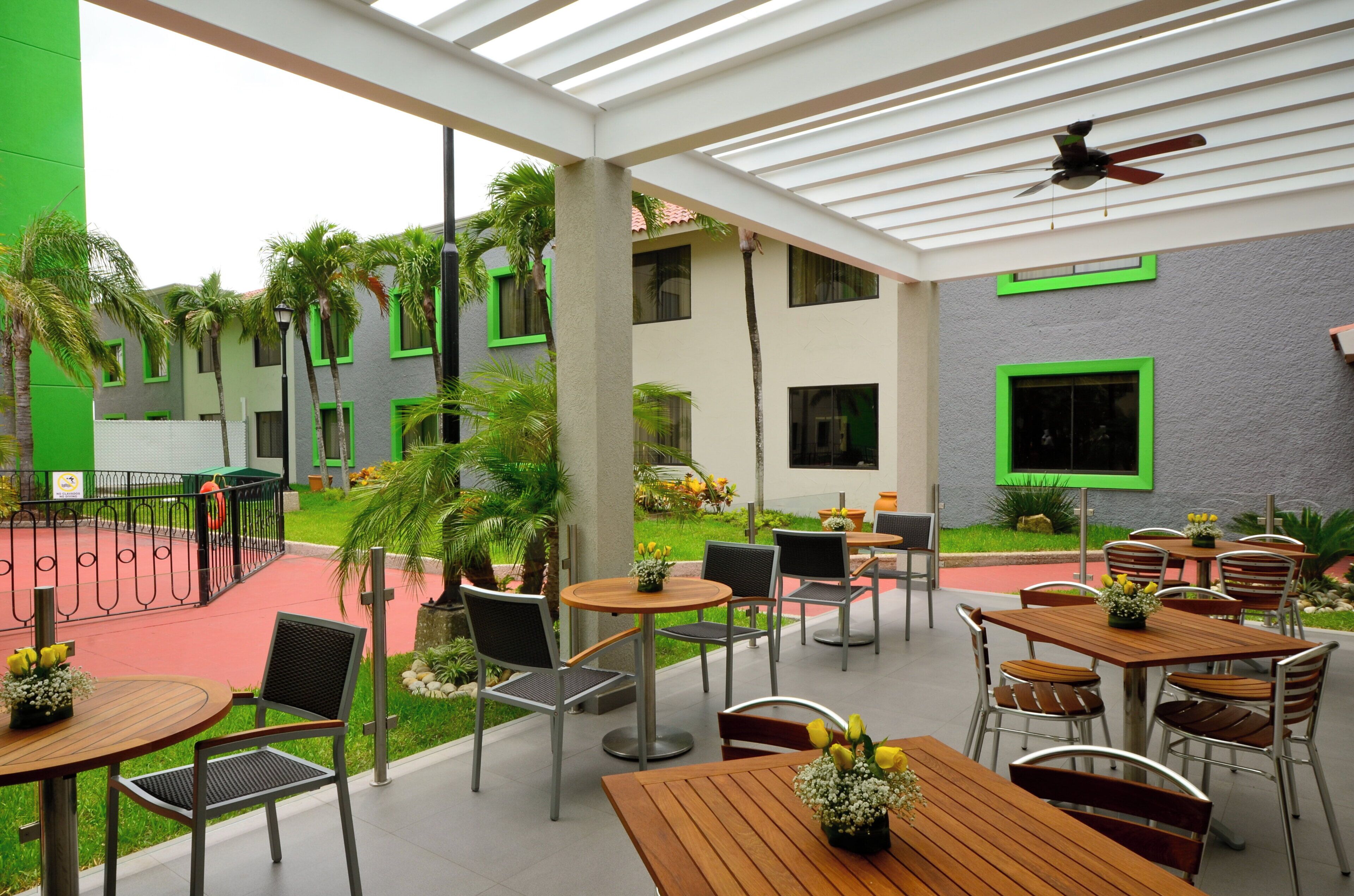Area patio esterna con diversi tavoli e sedie, circondata dal verde e da un edificio con finestre a cornice verde.