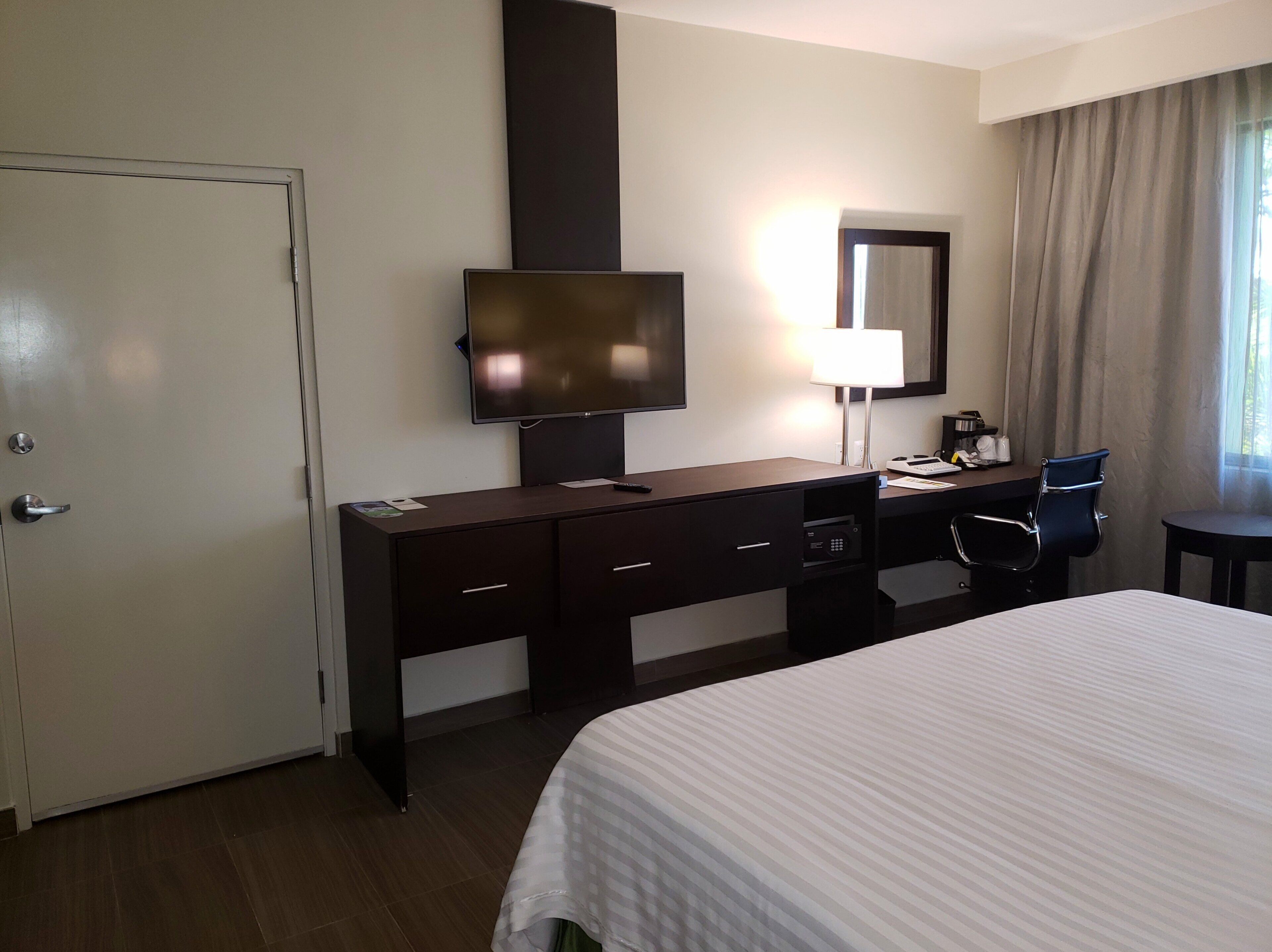 A hotel room with a bed, a desk with a chair, a television, a lamp, and a door.