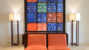 A lobby with two orange chairs, a coffee table with a book and a vase of flowers, and a wall with stacked shipping containers.