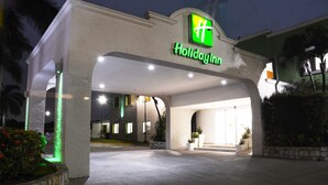 A Holiday Inn with a prominent green logo, a well-lit entrance, and a stone planter with red flowers.