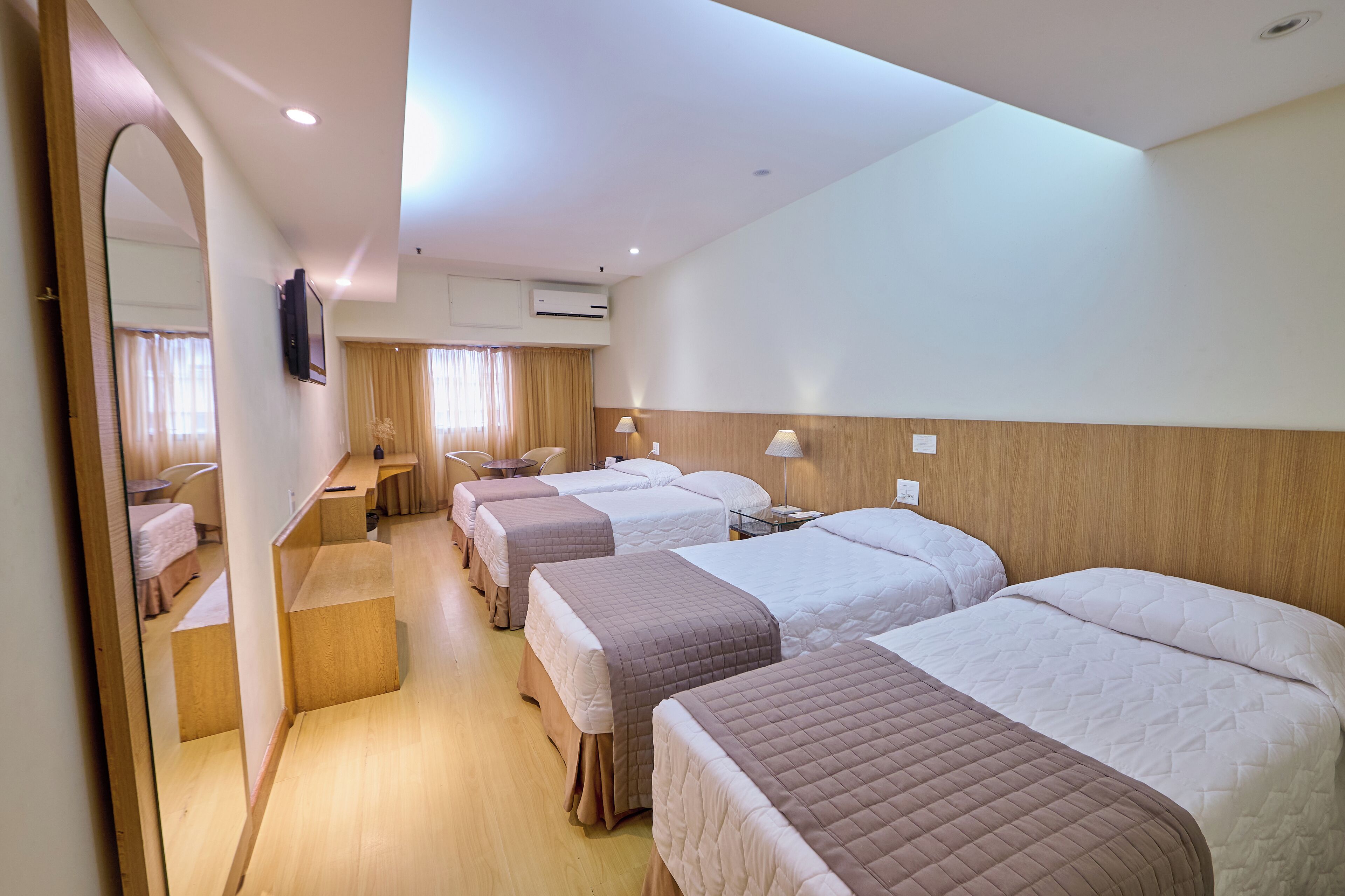 Une chambre d’hôtel avec quatre lits disposés en rangée, une tête de lit en bois, un bureau avec une chaise et un miroir.
