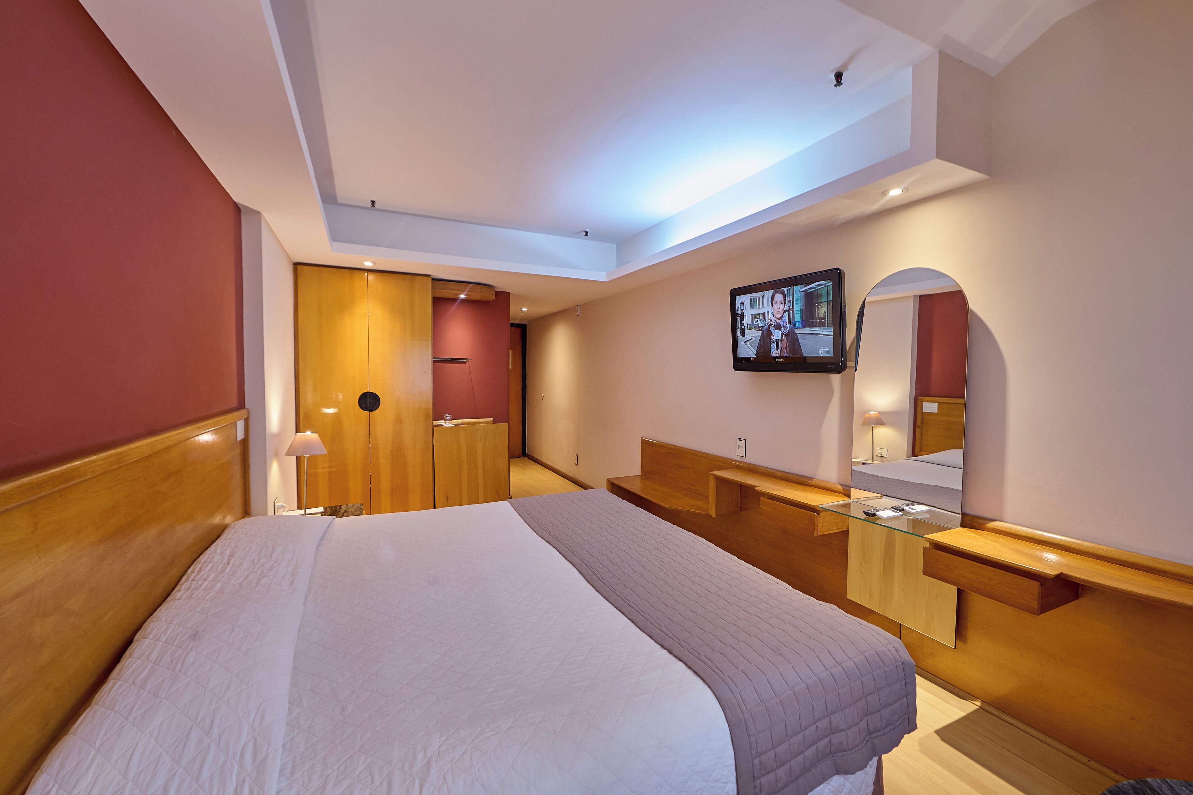 A modern hotel room with a large bed, a wooden headboard, a glass bedside table, a mirror, and a television mounted on the wall.