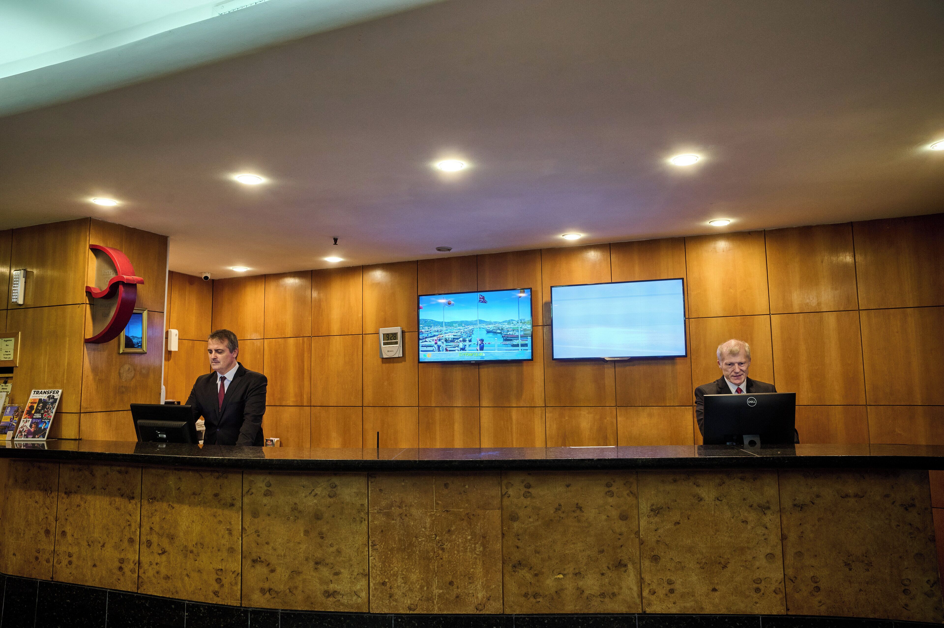 Un taulell de recepció amb dos empleats, panells de fusta i dos monitors penjats a la paret.