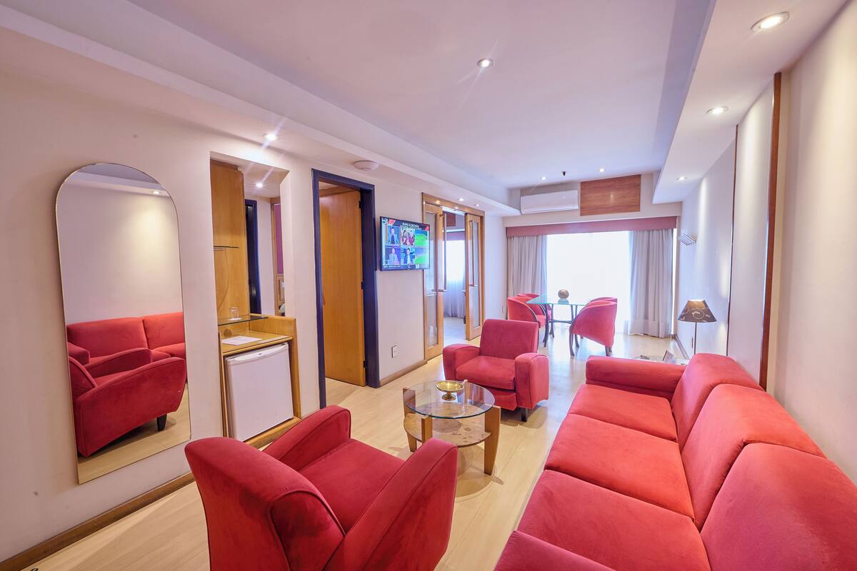 A modern hotel room with a red sofa set, a glass coffee table, and a large mirror. There is a TV mounted on the wall and a door leading to another room.