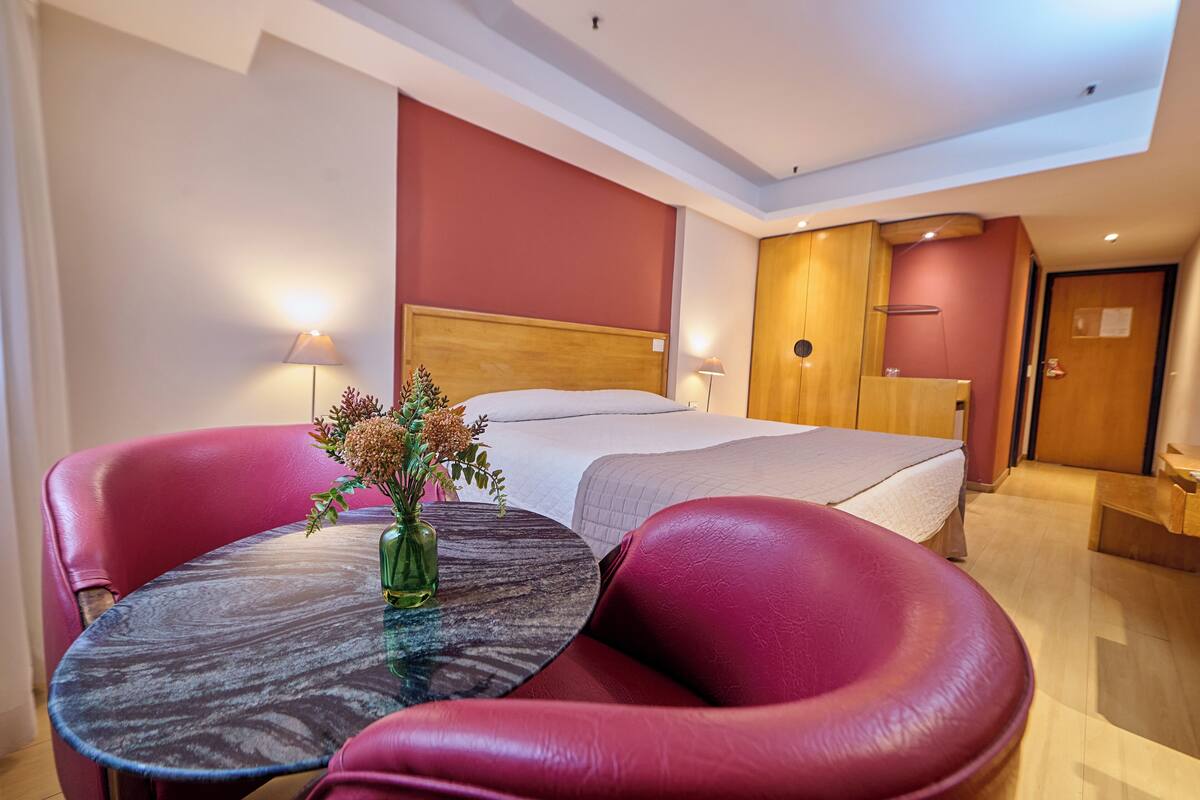 A hotel room with a bed, a round table with a vase, and two red chairs.